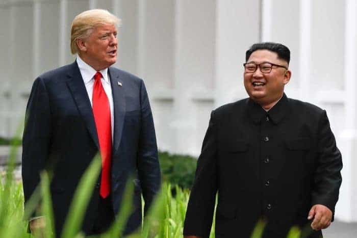 Donald Trump and Kim Jong-un smiling and walking along a grassy pathway.