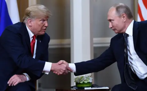 Getty Images Russian President Vladimir Putin and US President Donald Trump shake hands before a meeting in Helsinki. They are both sitting down, wearing dark suits and Donald Trump is saying something to Putin.