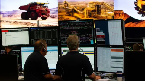 Rio Tinto Two men sit at a desk with multiple screens monitoring trucks and other mining equipment