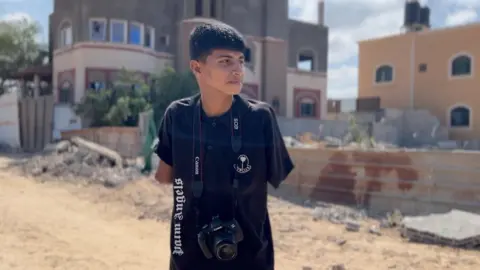 BBC Diya al-Adini stands in an empty Gaza street, with both arms missing and one stump visible underneath a black t-shirt and a camera hanging around his neck