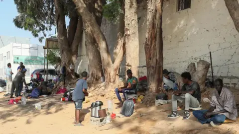 Getty Images Refugees from Sudan sit down in front of the UNHCR offices in Tripoli, Libya, on 15 July 2023. No-one pictured is in the article.