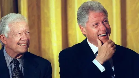 Getty Images US President Bill Clinton smiles as he listens to speakers with former US President Jimmy Carter in front of a yellow curtain during a rally in 2000 in support of permanent normal trade relations (PNTR) with China. 