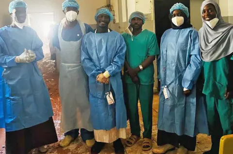 Mudathir Ibrahim Suleiman A group of medics at Saudi Hospital in el-Fasher in scrubs smile as they stand for a group photo after successfully performing two-hour emergency caesarean under bombardment. Dust from the shell damage can be seen on the floor