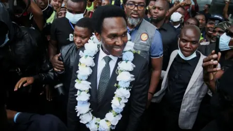 Reuters Venancio Mondlane comes outside the terminal building at the Maputo International Airport wearing a white garland around his next and surrounded by journalists and supporters.