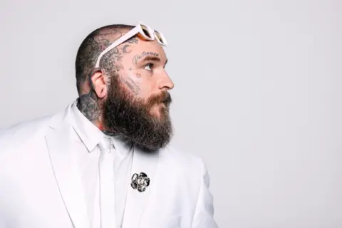 Claire Marie Vogel Teddy Swims, dressed in a white tux, poses sideways-on, with a pair of white glasses perched on his tattooed head